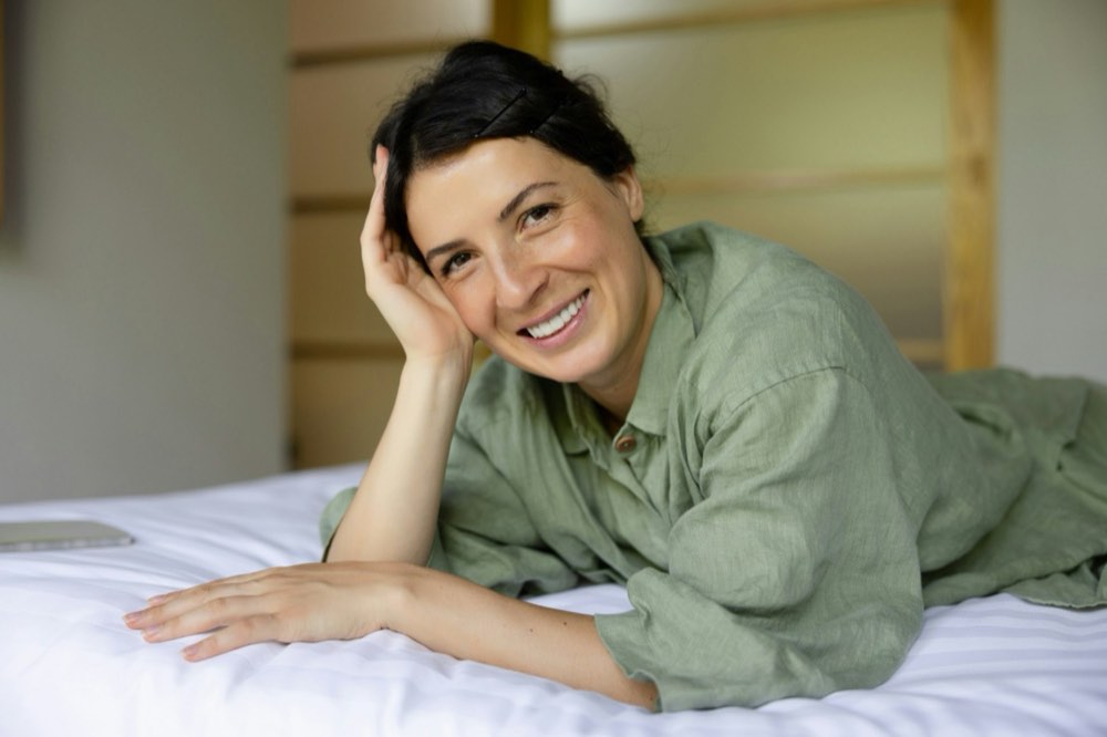 A woman lying on a bed in a light bedroom, resting on one arm