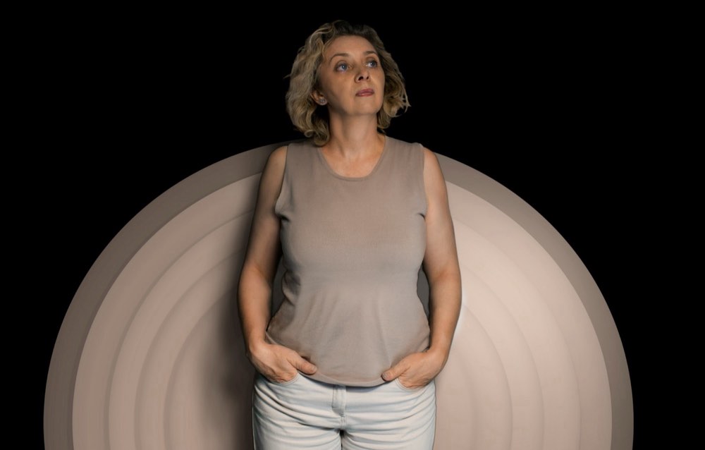 A woman standing against a dark background lit by concentric rings of warm light